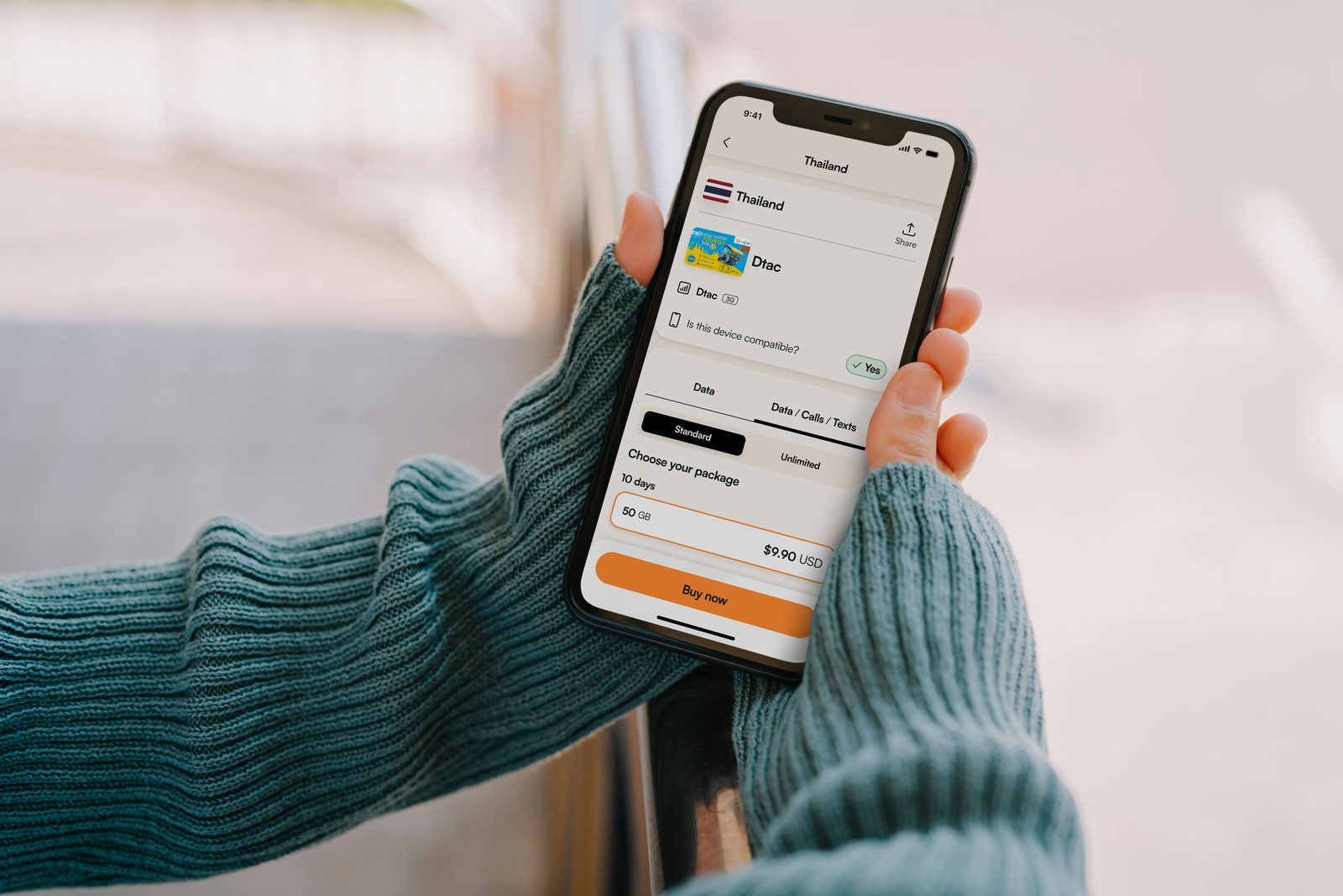 A person in a teal sweater holding a phone showing a mobile plan checkout screen with an orange Buy Now button