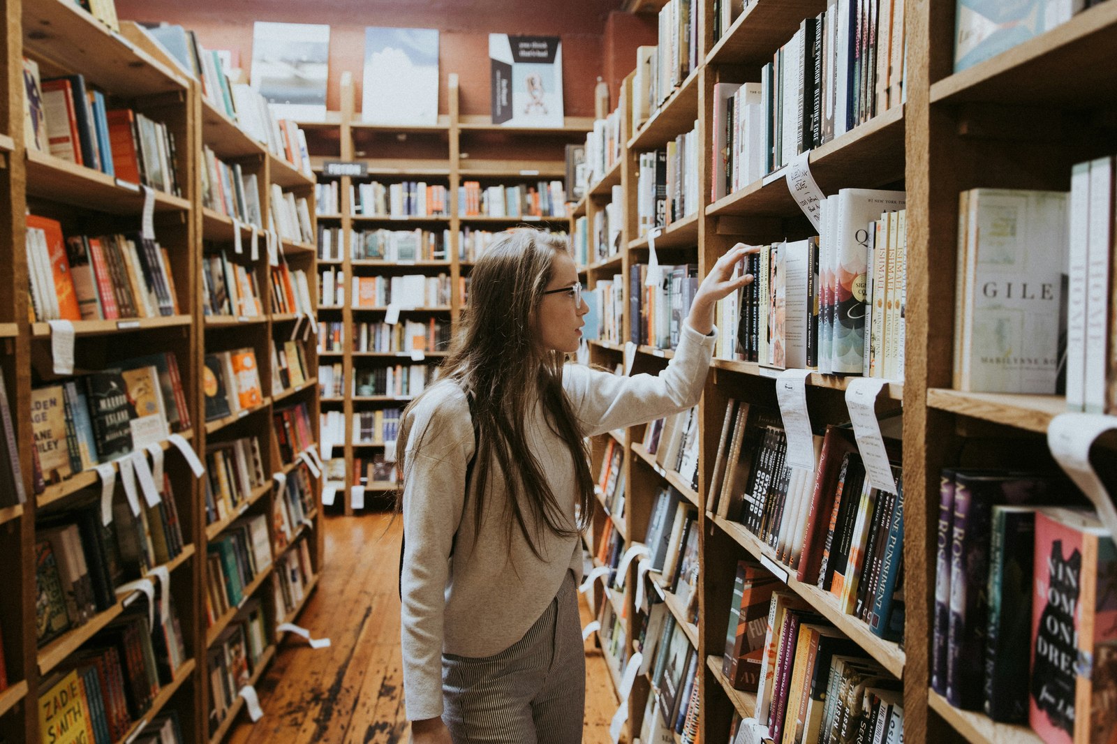 A woman browsing bookshelves in a bookstore, reaching up to pull a book from a high shelf