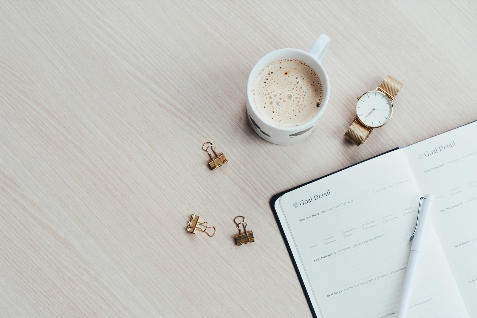 A planner open to a 'Goal Detail' page next to a cup of coffee, a gold watch, gold binder clips, and a pen