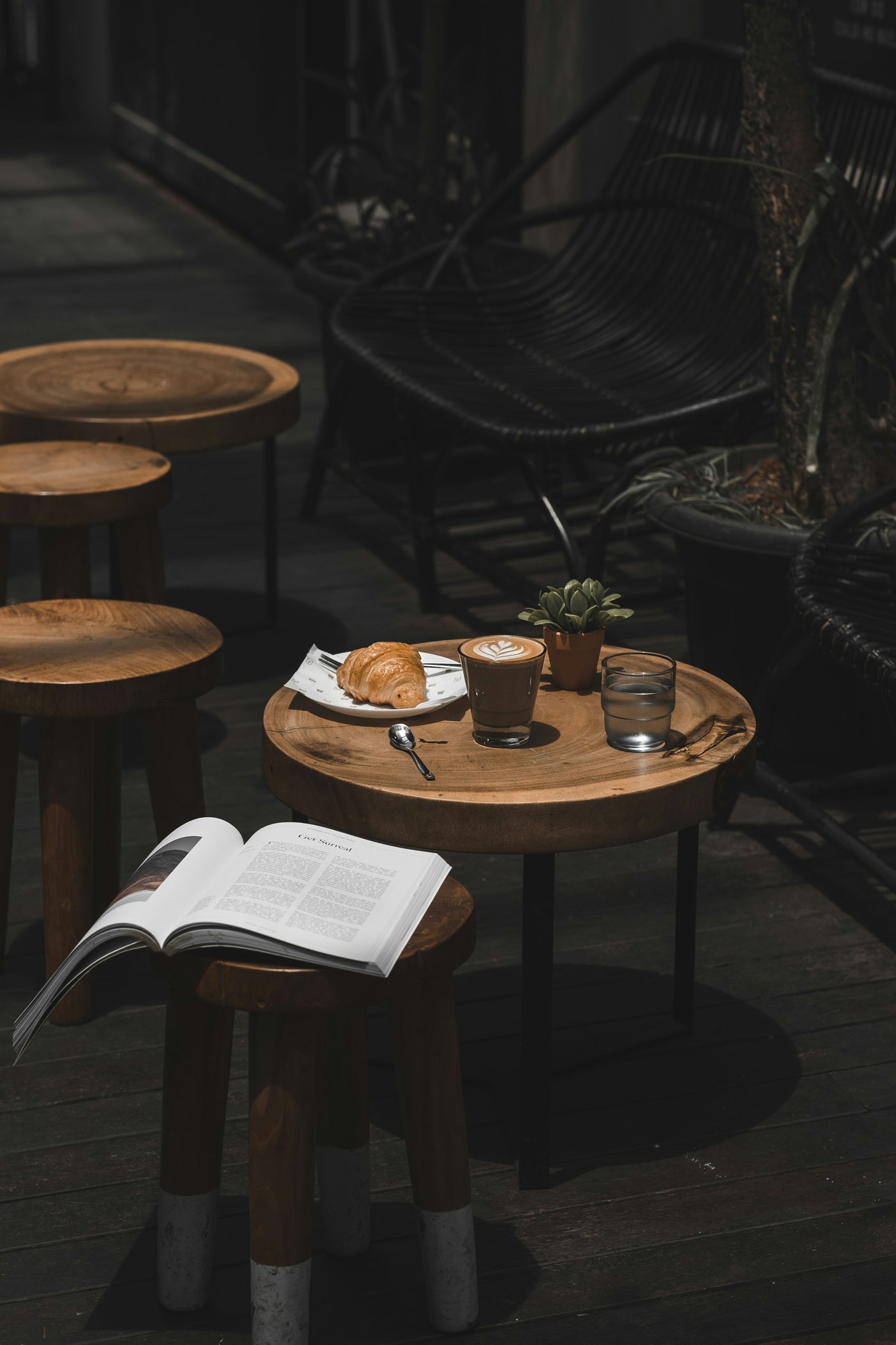 A croissant, latte with leaf-pattern foam, and glass of water on a round wooden cafe table, with an open magazine on a stool in the foreground
