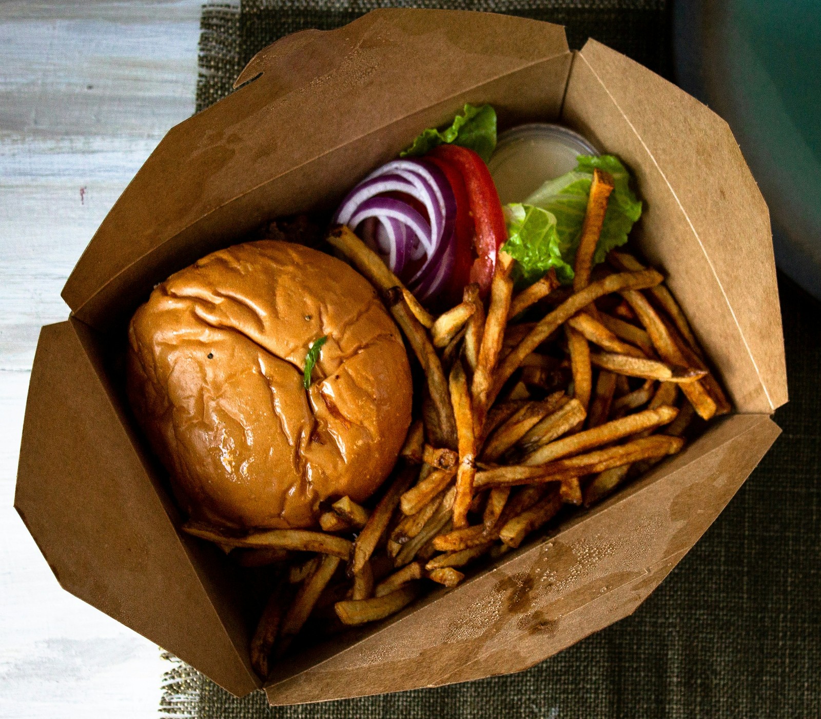 An overhead view of a takeout burger and fries in an open cardboard container, with lettuce, tomato, and red onion on the side