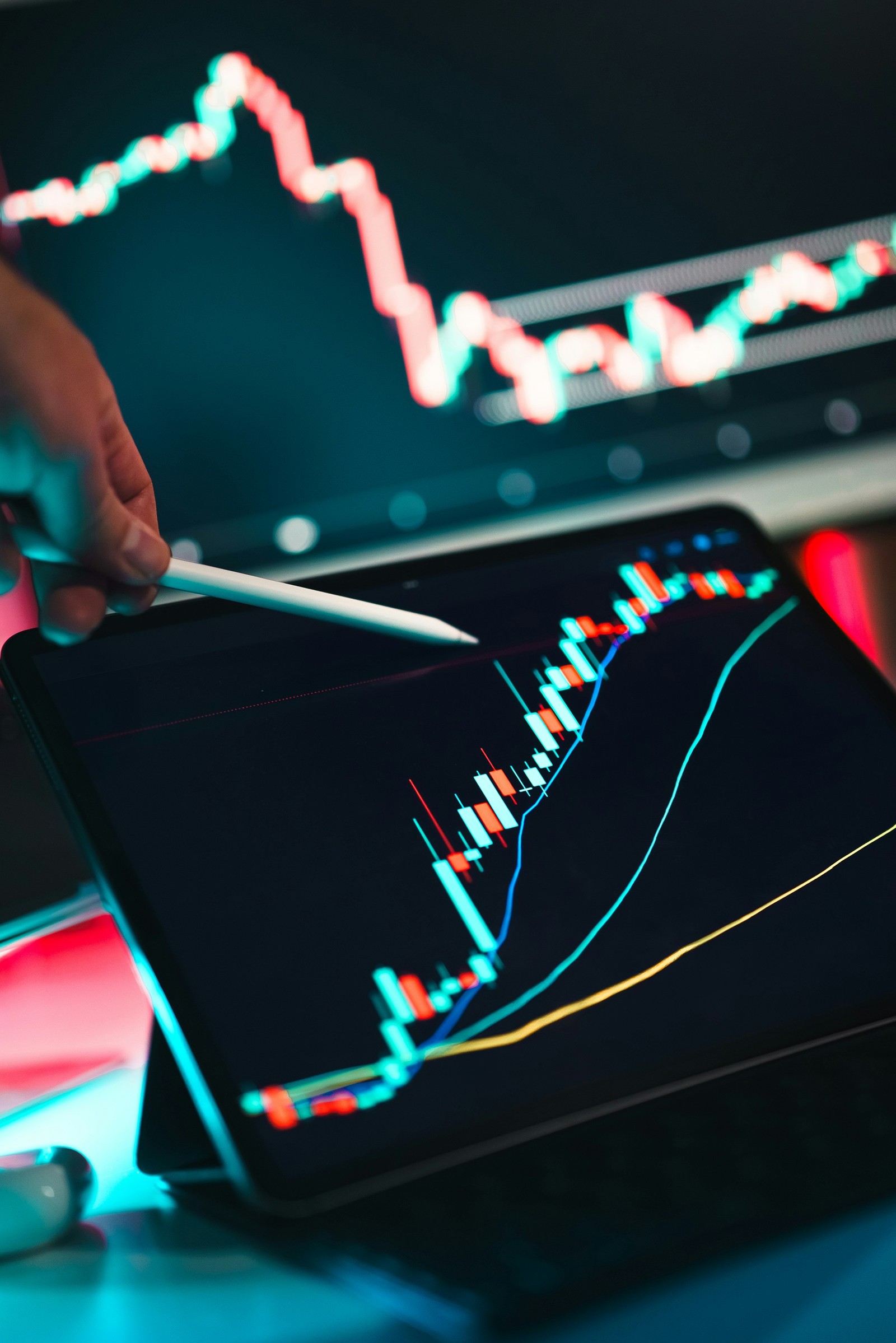 A hand holding a stylus pointing at a candlestick stock chart on a tablet screen, with a blurred stock ticker display glowing in the background